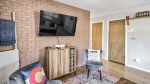 A living room with a brick wall, a flat-screen TV mounted above a wooden cabinet, a grey armchair with a cushion, and a patterned rug. - 5BR 💖heart Of Old Colorado Cityfoosball 💖 (Colorado Springs)