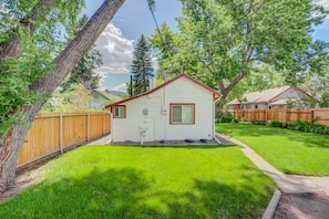 A small, single-story house with a red roof and white siding, surrounded by a wooden fence and lush green grass. - 1bdcute Tiny Housenear Olympic Training Center (Colorado Springs)