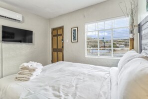 Une chambre à coucher avec un lit, un téléviseur fixé au mur, une fenêtre avec des stores et une porte.