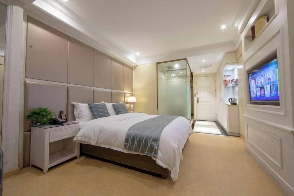 Quarto com cama, mesa de cabeceira, luminária, televisão e vista para um corredor.