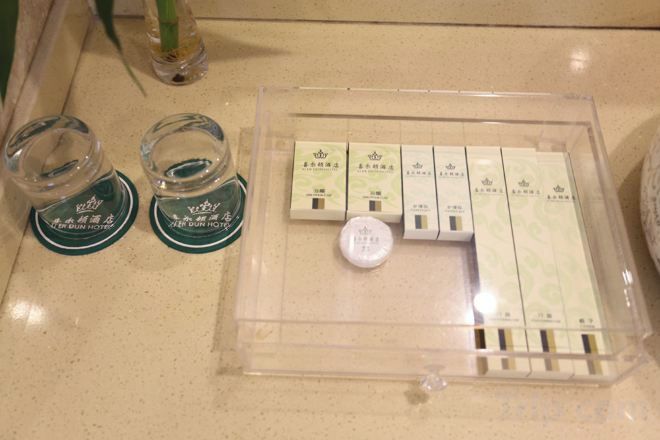 Embalagem transparente com vários produtos de higiene pessoal e uma garrafa de água sobre uma bancada.