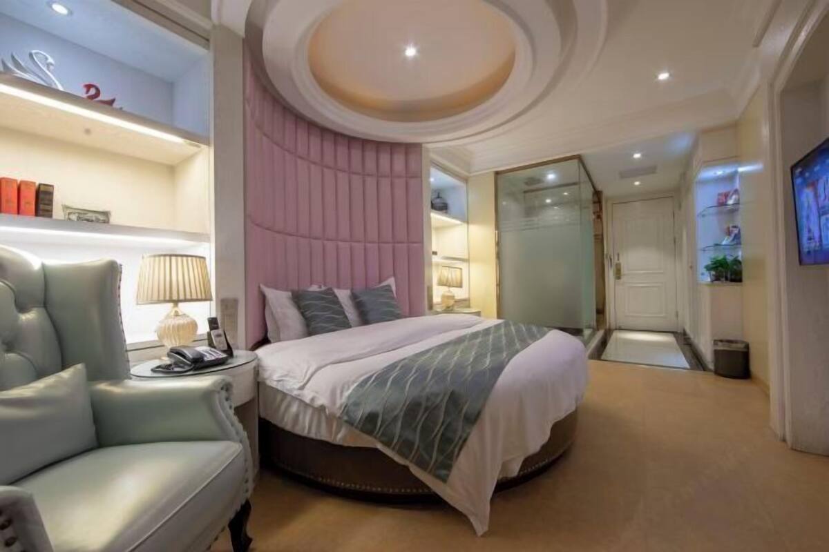 Um quarto moderno com uma cama redonda, um chaise lounge, uma parede em tons de rosa e uma TV fixada na parede.