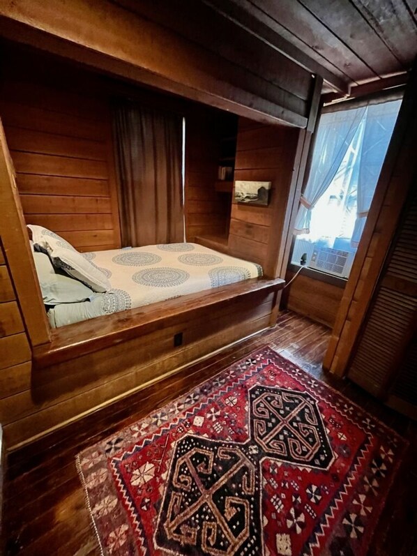 A wooden cabin room with a bed, a window with curtains, and a patterned rug.
