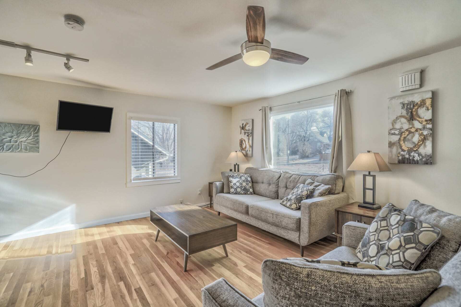 A spacious living room with a ceiling fan, a large window with blinds, a sofa set, a coffee table, and a TV mounted on the wall.