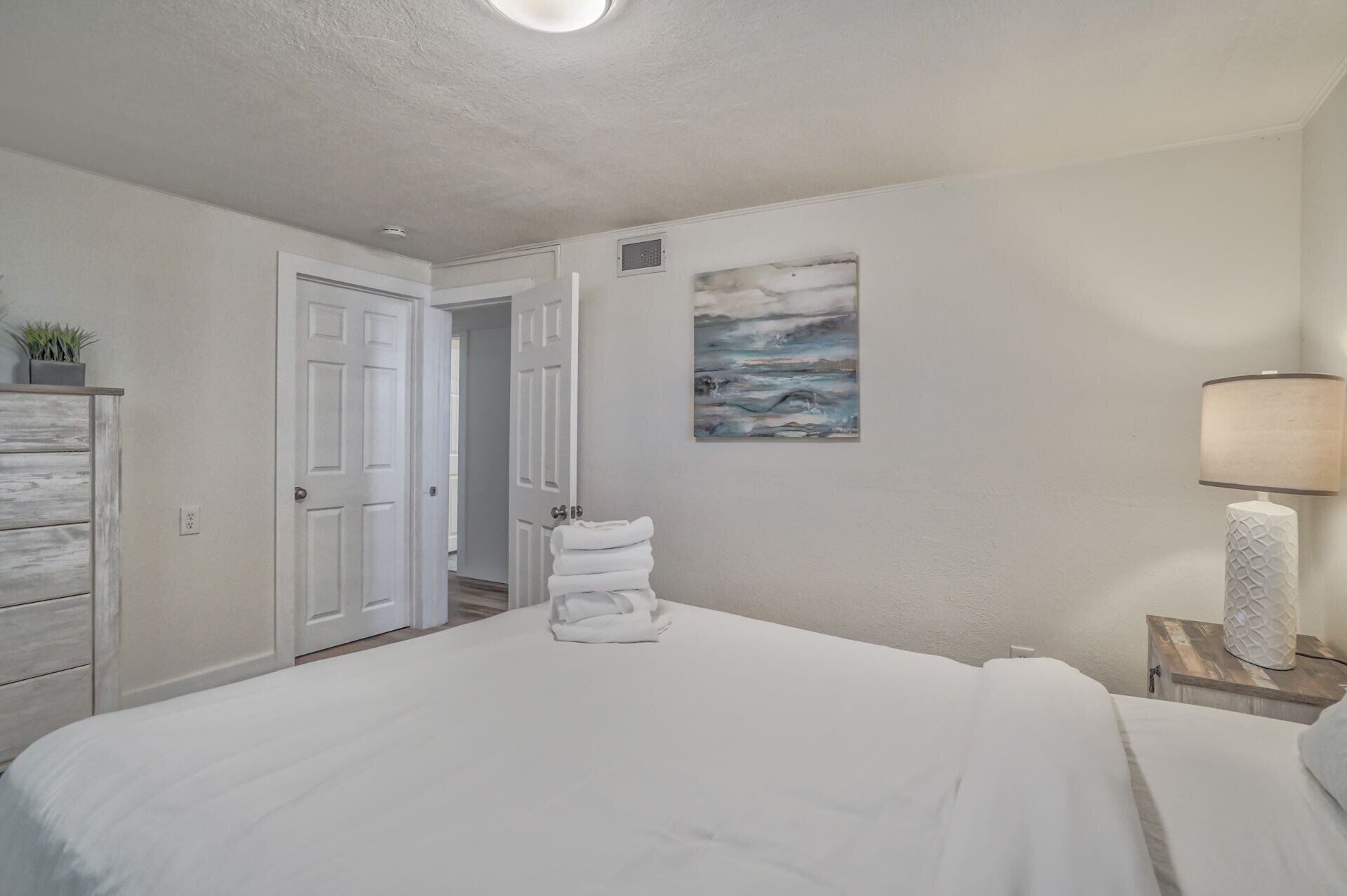 Ein Schlafzimmer mit einem großen Bett, einem Bild an der Wand, einer Lampe und einer Tür zu einem anderen Raum.