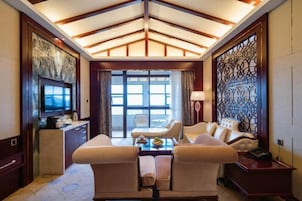 Una habitación de hotel espaciosa con un ventanal grande, un televisor de pantalla plana, un área de comedor con mesa y sillas, y un panel decorativo en la pared.