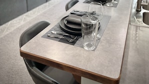 Moderni keittiö, jossa on pitkä ruokapöytä ja neljä tuolia, sekä suuret ikkunat, joista avautuu näkymä maaseudulle.