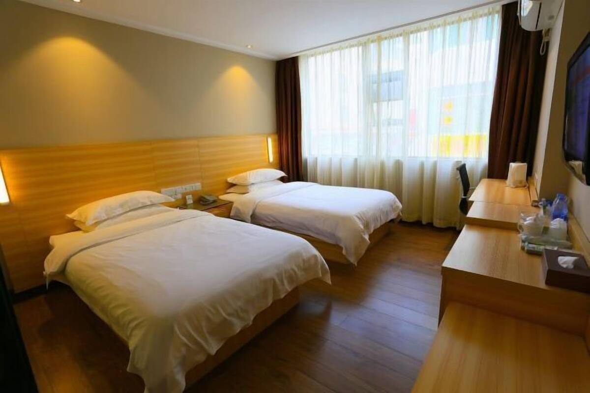 A hotel room with two beds, a wooden headboard, a desk with a chair, and a large window with curtains.