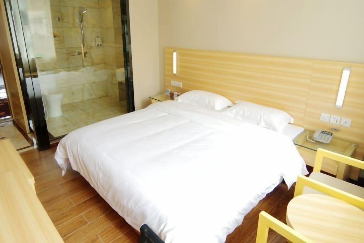 Una habitación de hotel con una cama grande, una mesita de noche con teléfono, cabina de ducha de vidrio y suelo de madera.