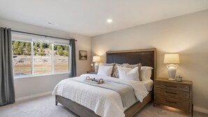 A bedroom with a large bed, two bedside tables, a window with curtains, and a view of a natural landscape.