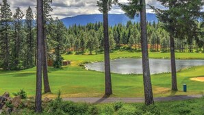A golf course with a pond, surrounded by tall trees and mountains in the background.