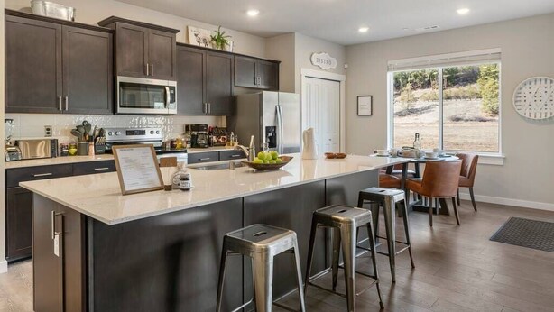 A modern kitchen with dark cabinets, a central island with stools, a dining area with a table and chairs, and a view through large windows.