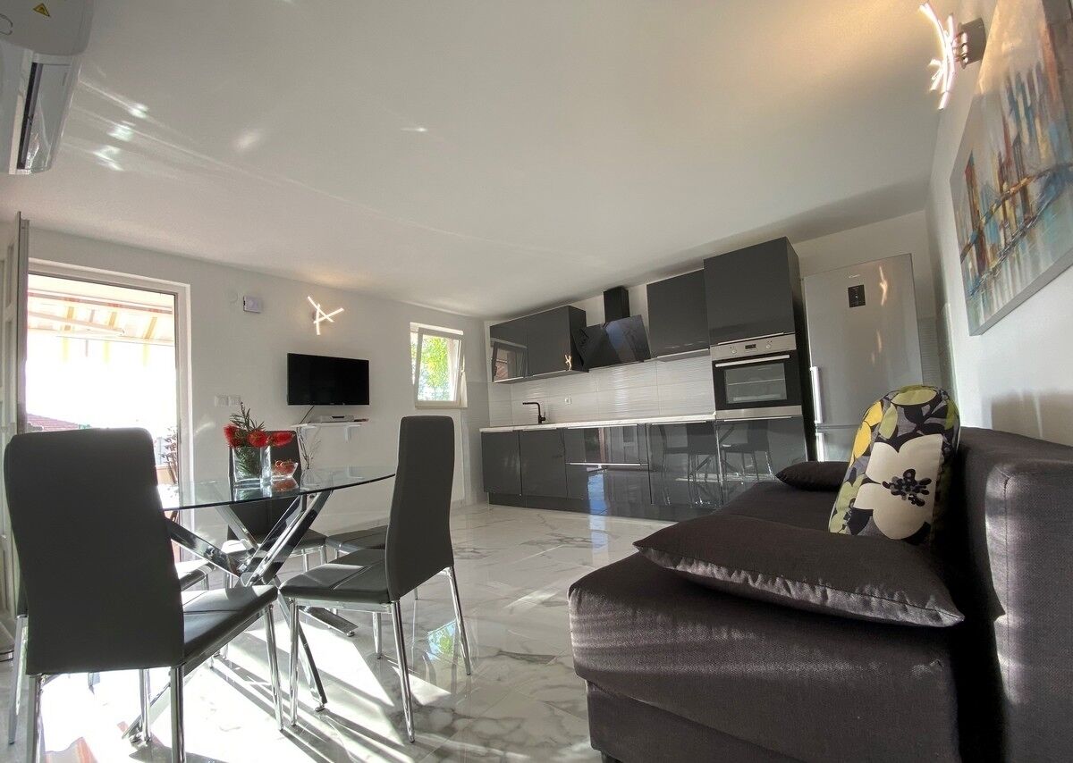 A modern kitchen with a flat-screen TV, a dining area with a glass table and chairs, and a living area with a sofa and a painting.