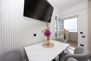 A modern room with a white marble table, grey chairs, a vase with purple flowers, and a large window offering a view of the sea.
