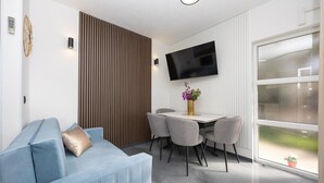 A modern living room with a blue sofa, a dining table with chairs, a flat-screen TV mounted on the wall, and a large window with a view of greenery. - Apartment in Okrug Gornji with Seaview, Air condition, WIFI, Washing machine (5049-1) (Okrug Gornji)