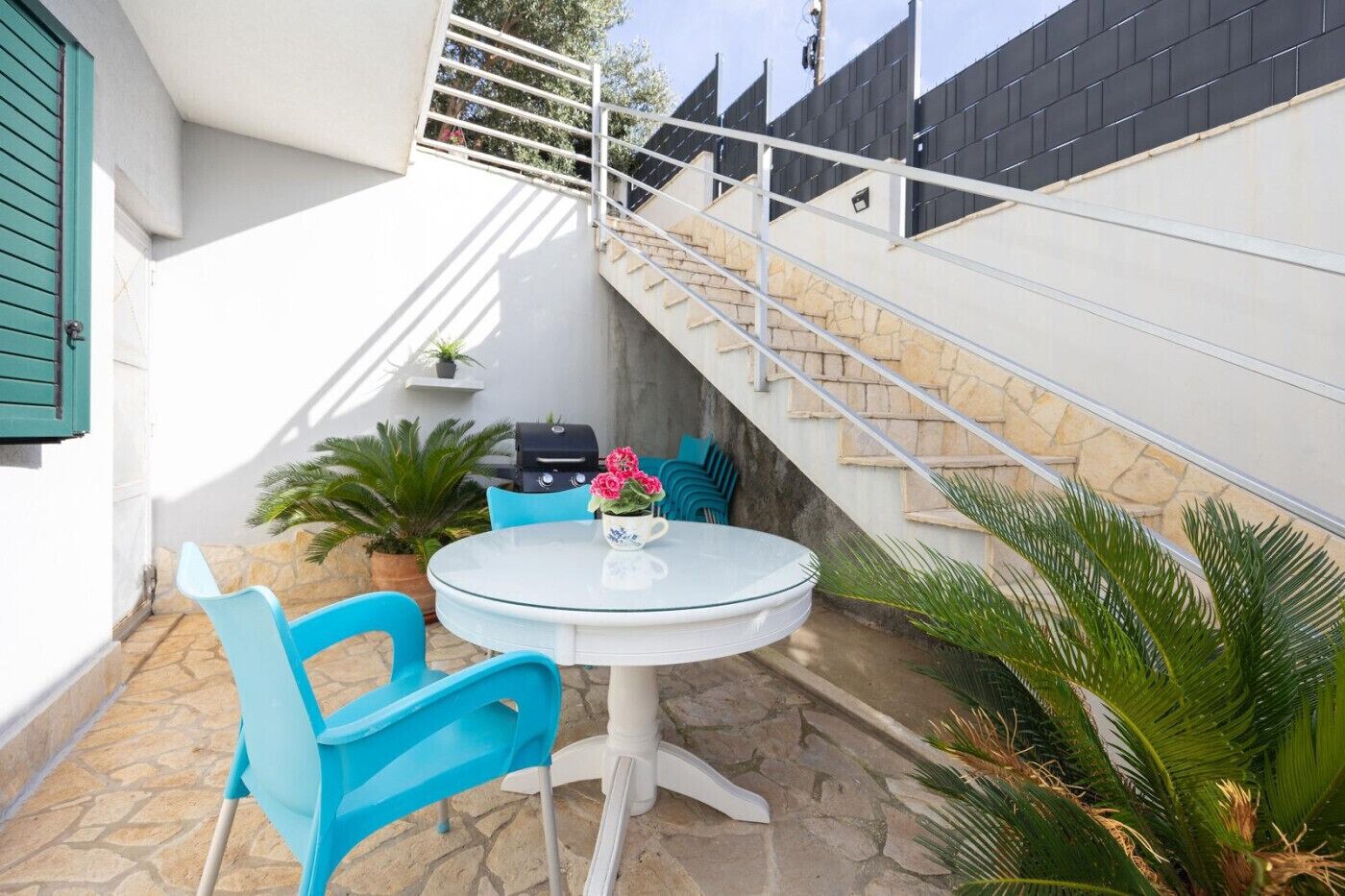 Une terrasse avec une table ronde et des chaises bleues, un barbecue et un escalier.