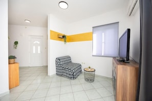 Una sala moderna con un sofá, una mesita redonda, un televisor y una ventana con persianas.