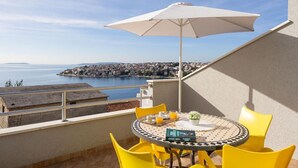 Un balcon avec une table et des chaises, un parasol blanc et une vue sur une ville côtière.