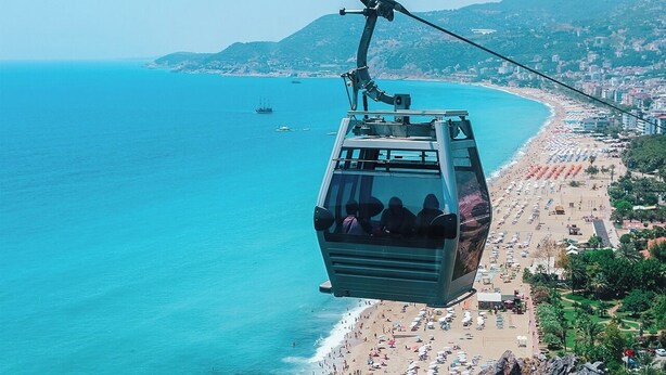 A cable car over a beach with clear blue water, a sandy shore, and a hilly landscape in the background. - Anjeliq Downtown Hotel (Alanya)