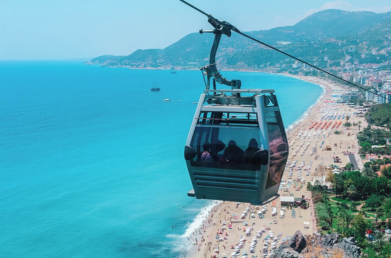 A cable car over a beach with clear blue water, a sandy shore, and a hilly landscape in the background.