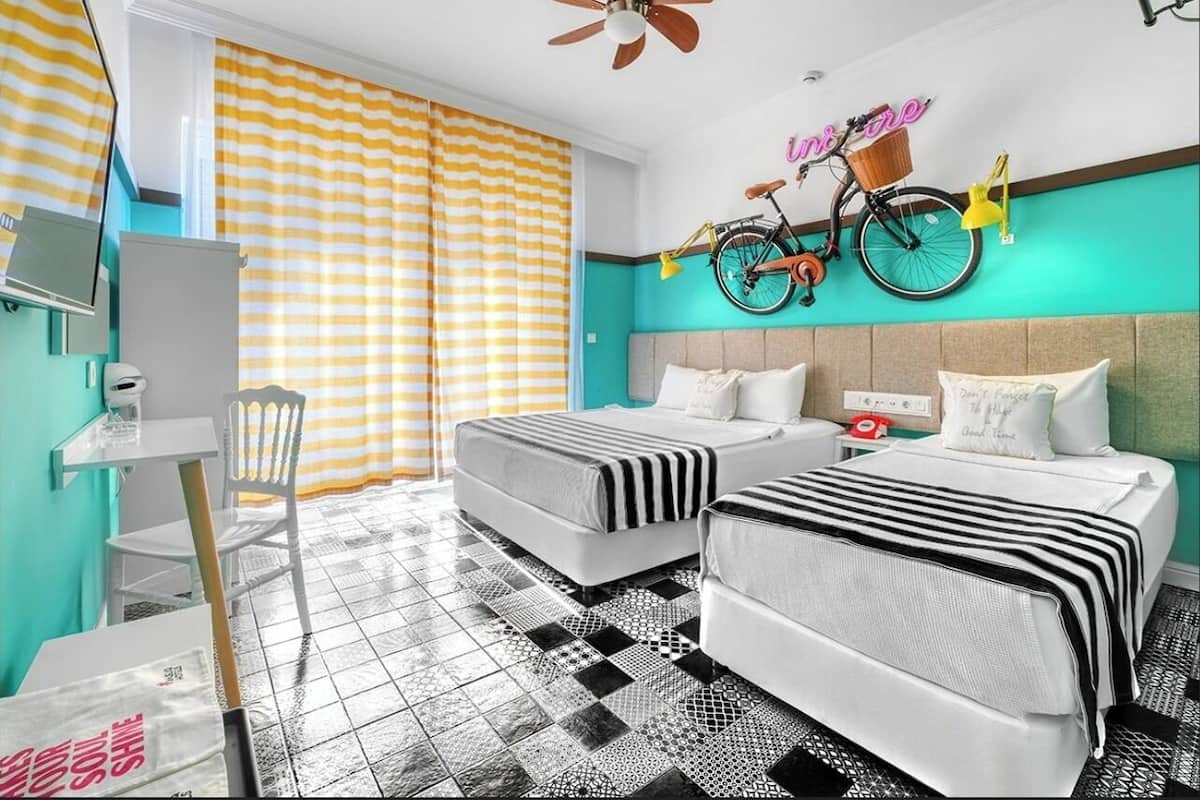 A hotel room with two beds, a desk, a chair, a bicycle, and a neon sign on the wall.