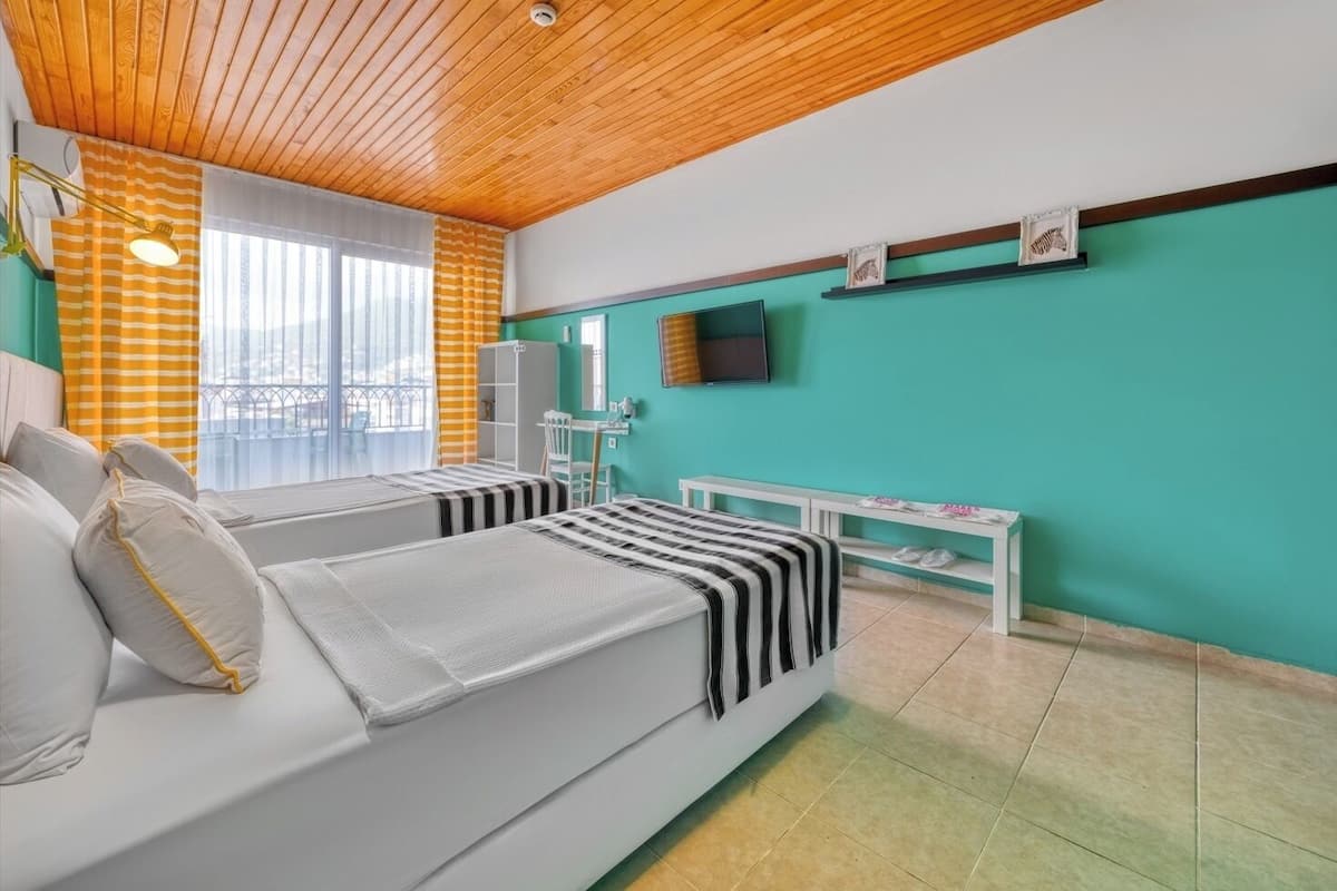A room with two beds, a TV, a window with curtains, a small table, and a shelf with decorative items.