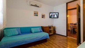 Une pièce avec un plancher en bois, un canapé bleu, un téléviseur fixé au mur et une porte qui donne sur une autre pièce.