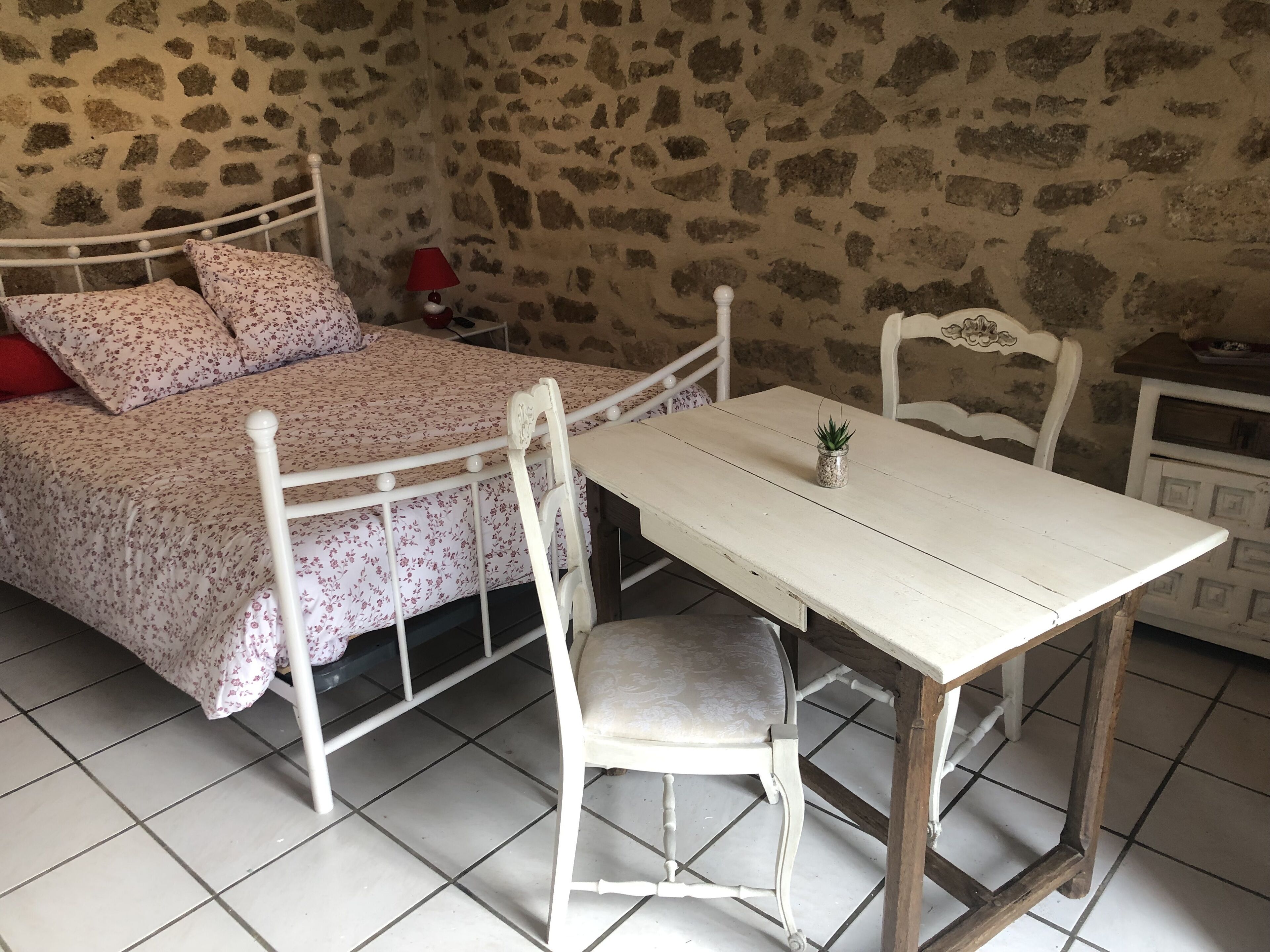 A bedroom with a bed, a table, and chairs. The room has stone walls and tiled flooring.