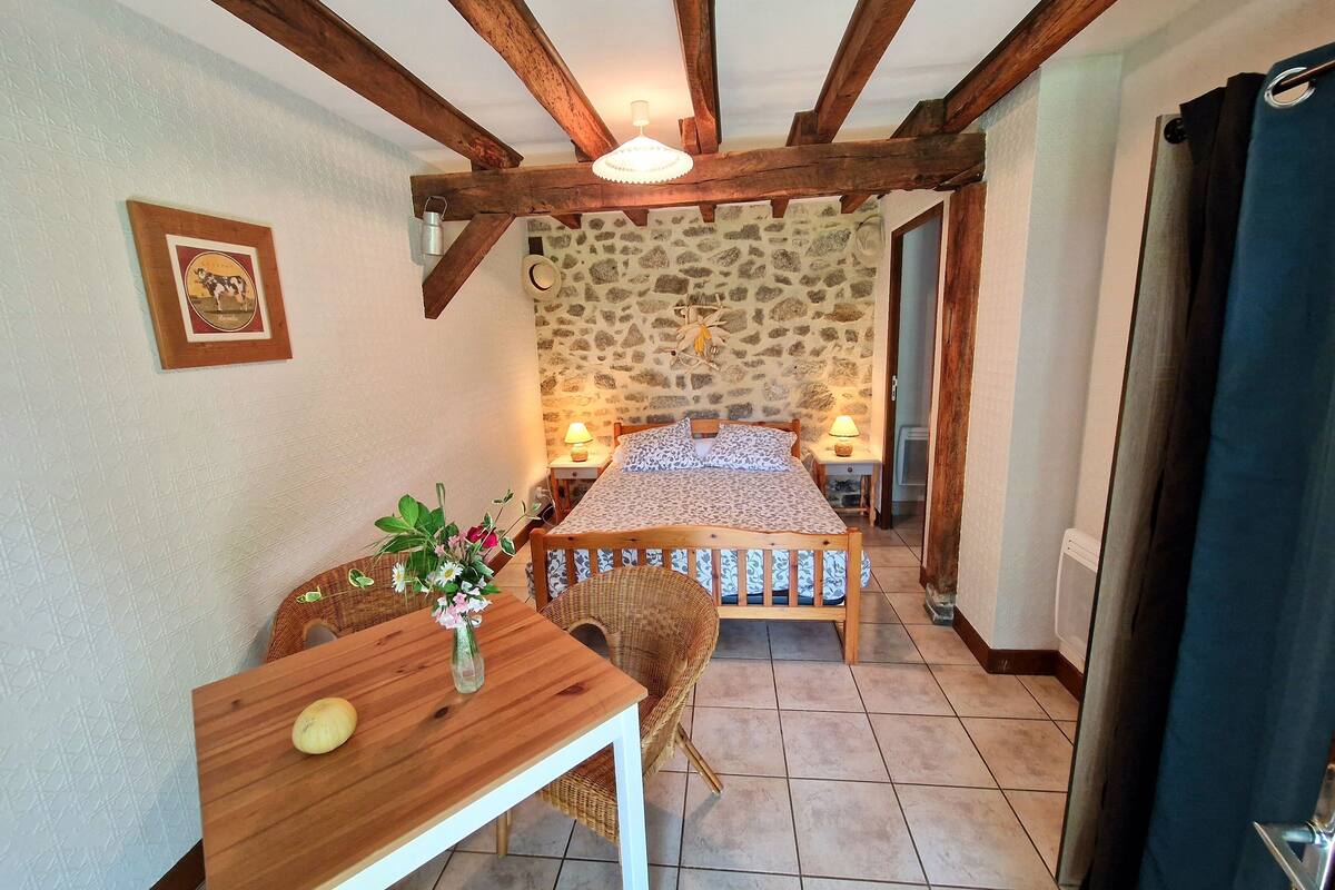 A cozy room with a bed, a wooden table with a vase of flowers, a wicker chair, and a stone wall.