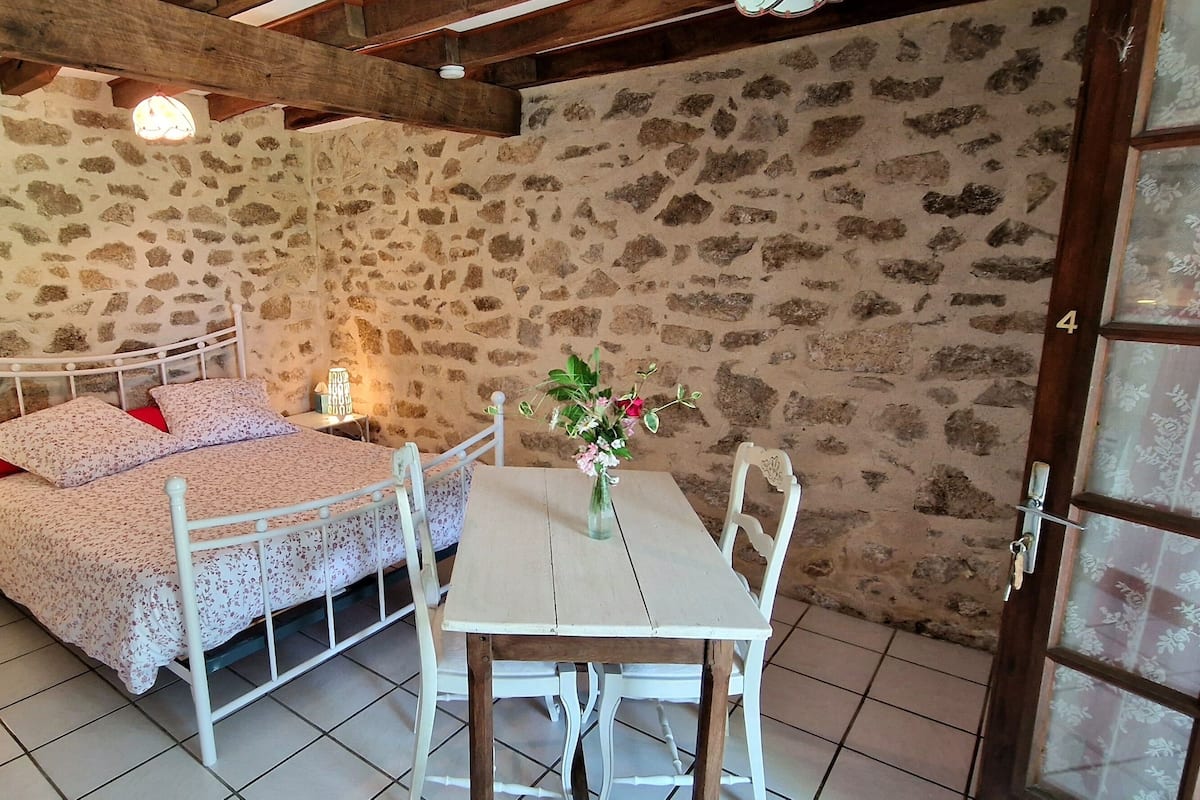 A room with a stone wall, a bed with floral bedding, a white table with chairs, and a small vase with flowers.