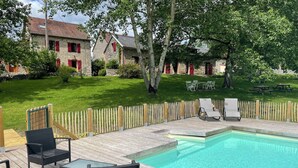 A wooden deck with a pool, outdoor furniture, and a stone building with red shutters in the background. - Maison d'hôtes Ourdeaux et gîte Chez Rouchon (Alleyrat)