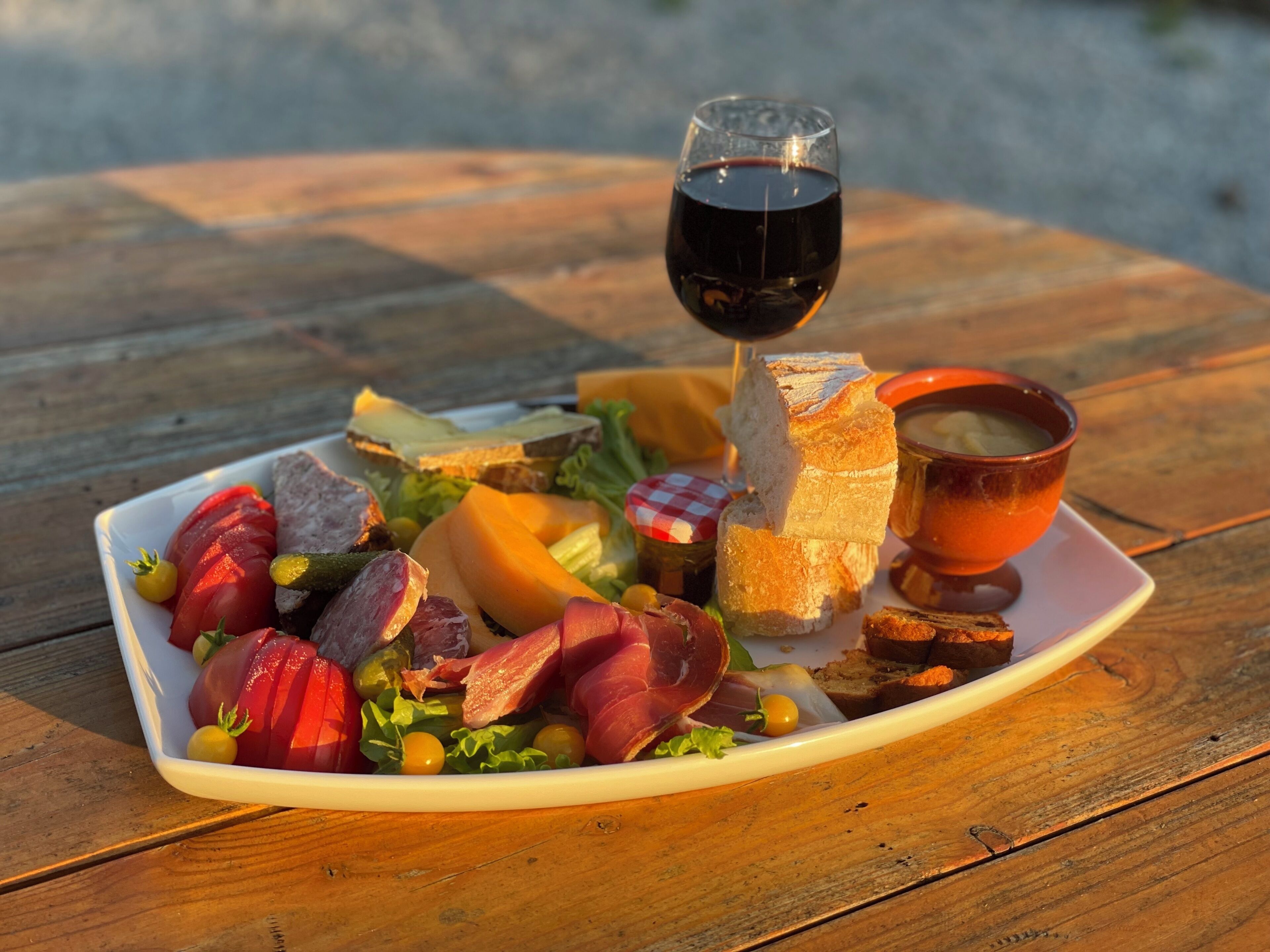 A square white plate with a variety of foods including sliced meats, cheese, and vegetables, accompanied by a glass of red wine and a bowl of soup on a wooden table.