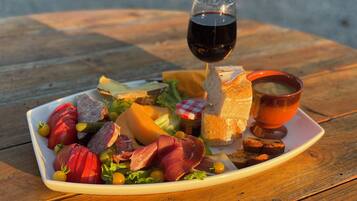 A square white plate with a variety of foods including sliced meats, cheese, and vegetables, accompanied by a glass of red wine and a bowl of soup on a wooden table.