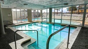 Una piscina interior con paredes de vidrio transparente, suelo de baldosas y vistas a la playa y palmeras fuera.