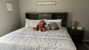 A neatly made bed with a patterned quilt, a teddy bear, and a bedside table with a lamp and a clock. - Adventure Acres- Suite C5 (Holly)