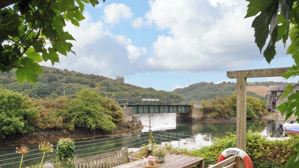 A wooden table and benches by a river with a bridge in the background. - Island House (Fowey)