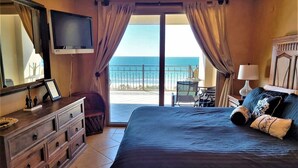 A bedroom with a large bed, a dresser, a TV, and a balcony with a view of the ocean.