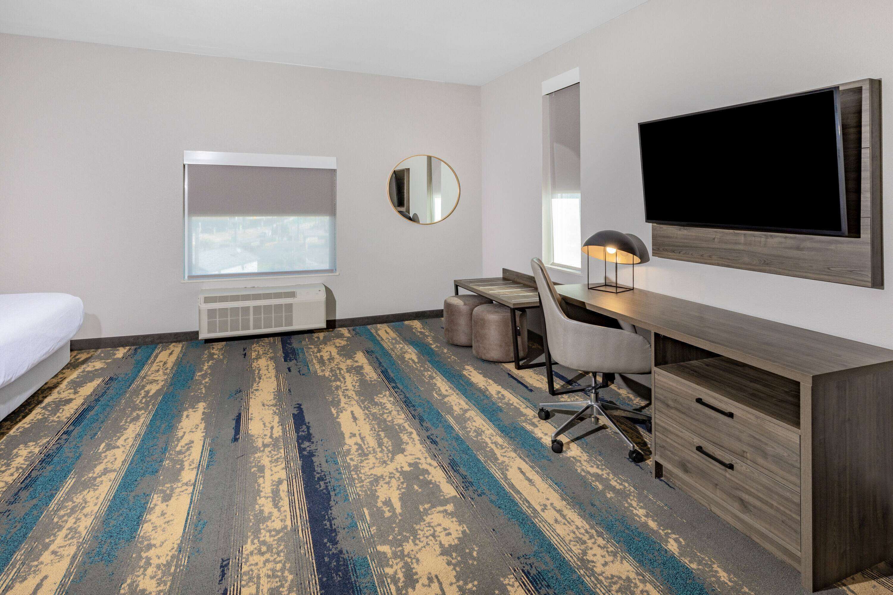 A hotel room with a bed, a desk, a chair, a large flat-screen TV, a round mirror, and a window with a view.