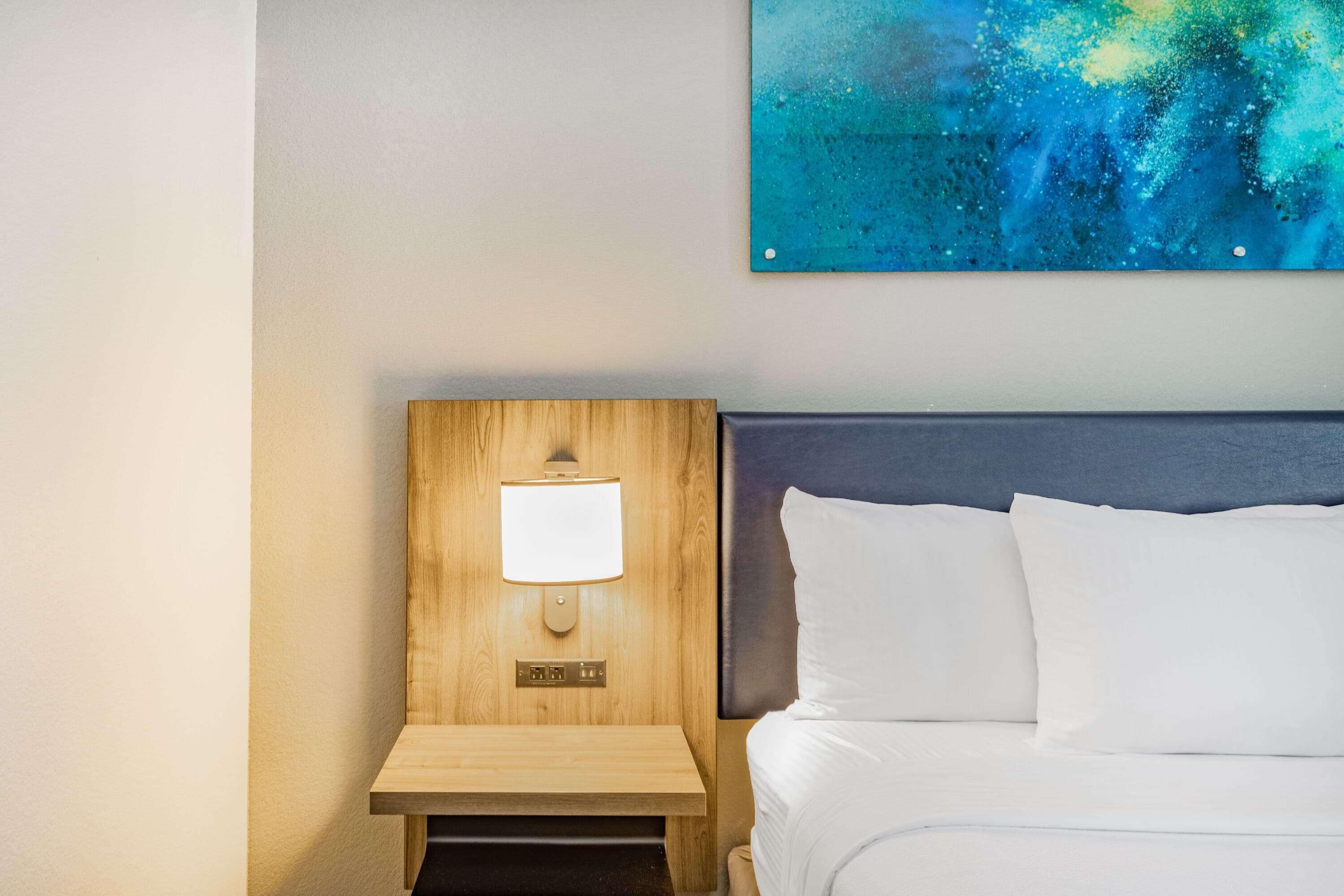 Hotelkamer met een houten nachtkastje, een nachttafel, een bed met witte lakens en een wandlamp.