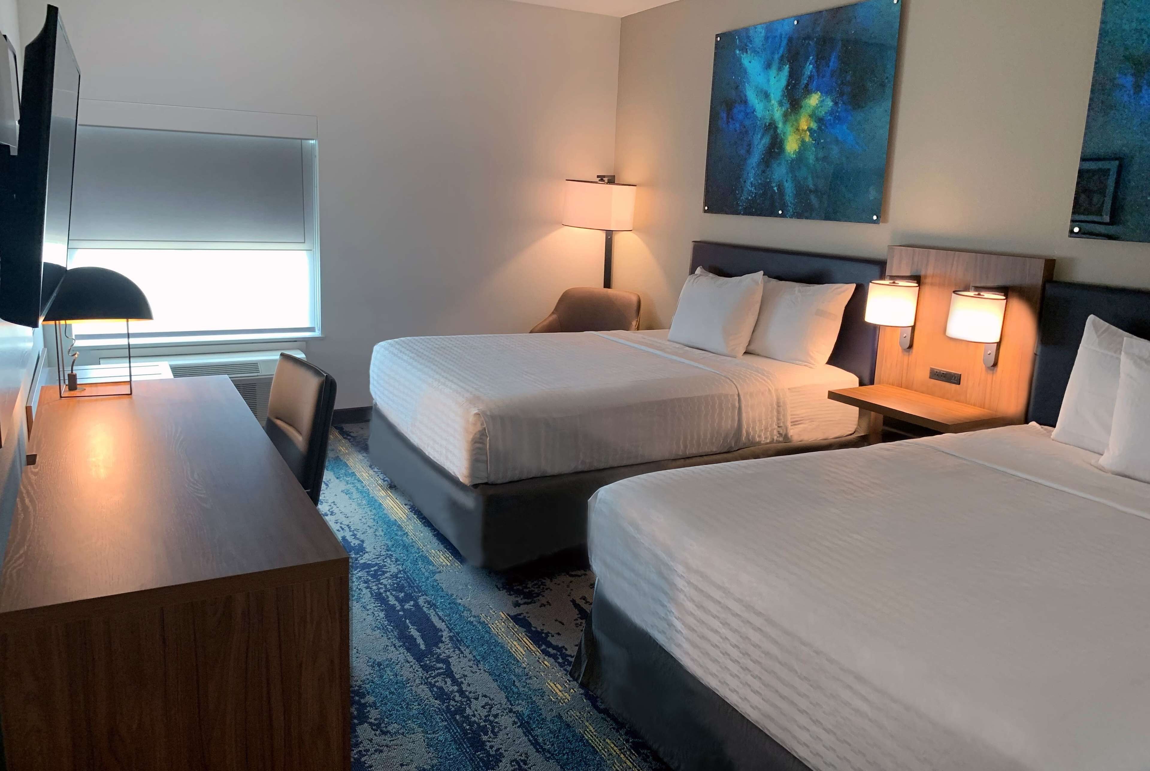 A hotel room with two beds, a desk, a chair, a TV, and a large painting on the wall.