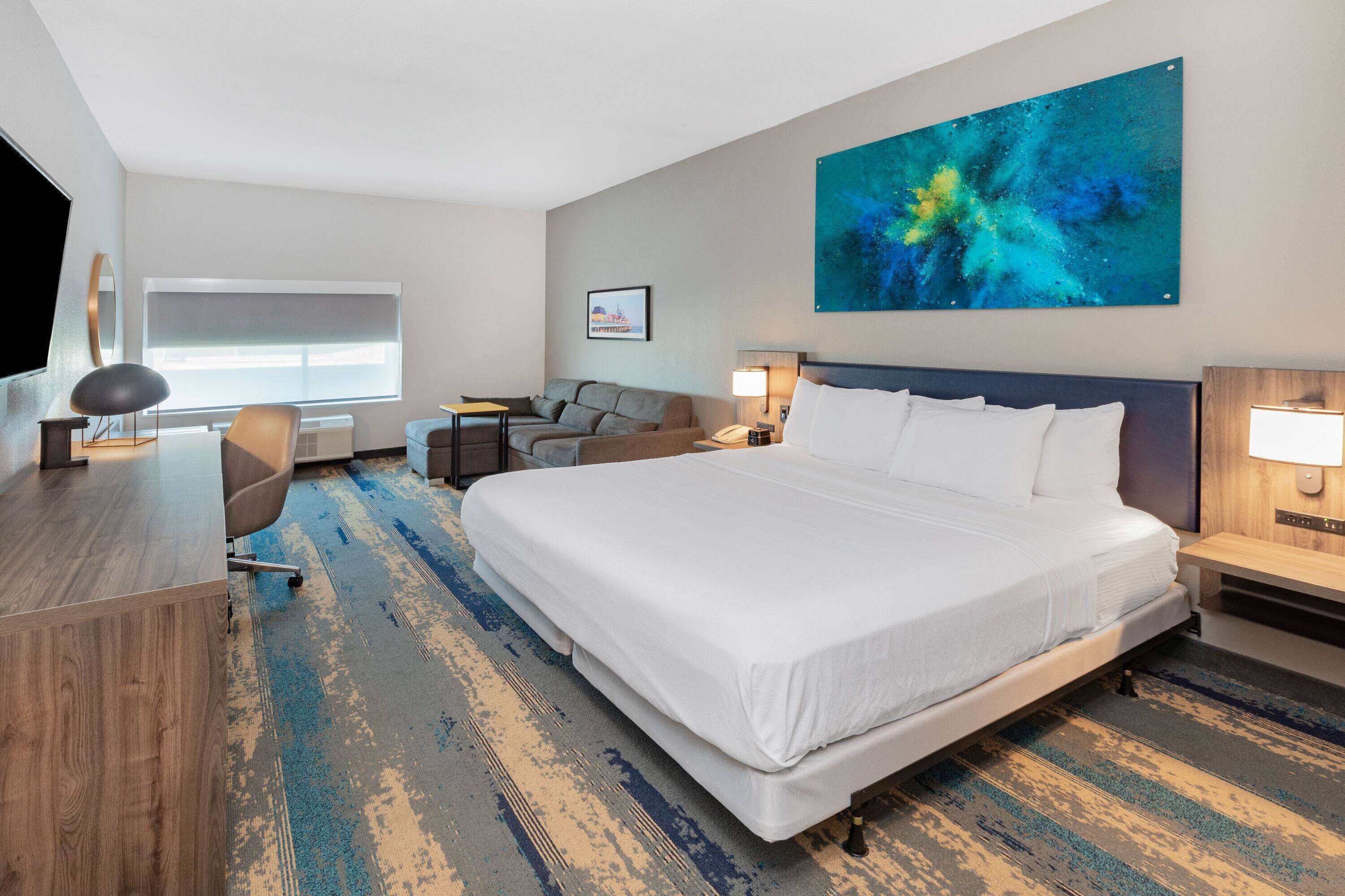 A hotel room with a large bed, a desk, a chair, a sofa, and a large painting on the wall.