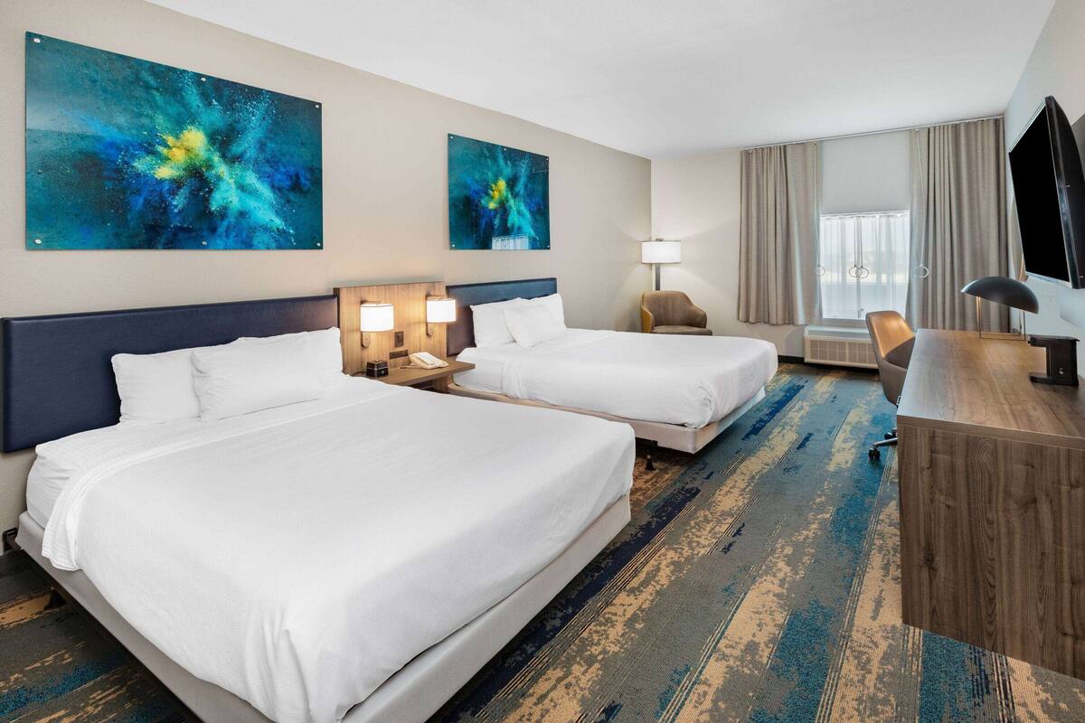 Une chambre d’hôtel avec deux lits, un bureau, une chaise, une télévision et des œuvres d’art accrochées aux murs.