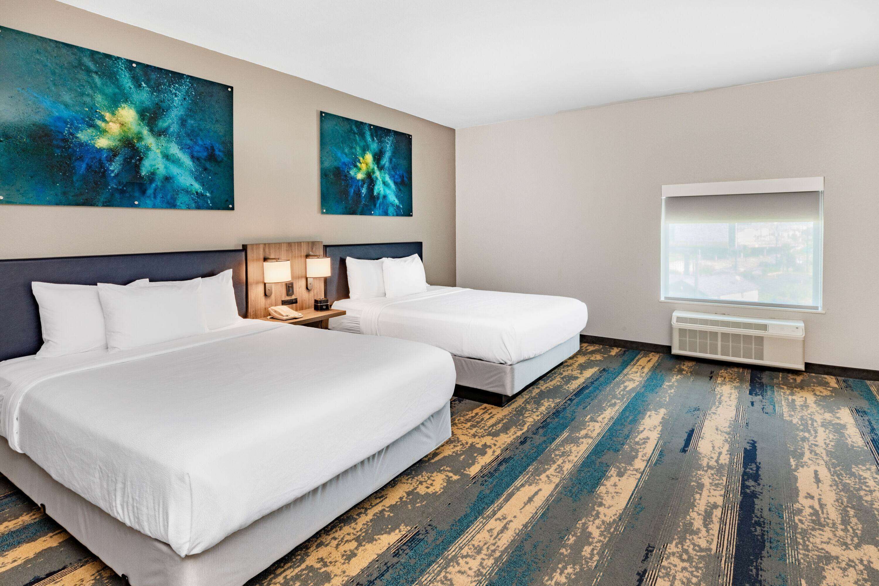 A hotel room with two beds, a large window, and abstract paintings on the wall.