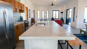 A modern kitchen with a central island, stainless steel refrigerator, wooden cabinets, and a large window providing natural light.