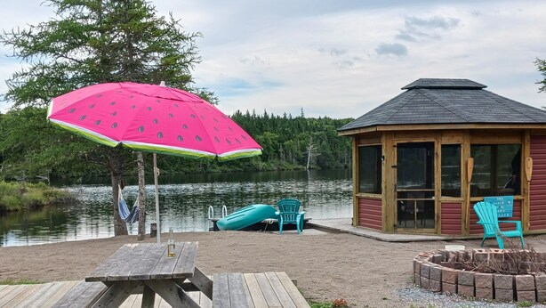 Une terrasse en bois avec une table de pique-nique, un parasol rose et une petite structure en bois sur laquelle est accrochée une pancarte indiquant « The Beach ».