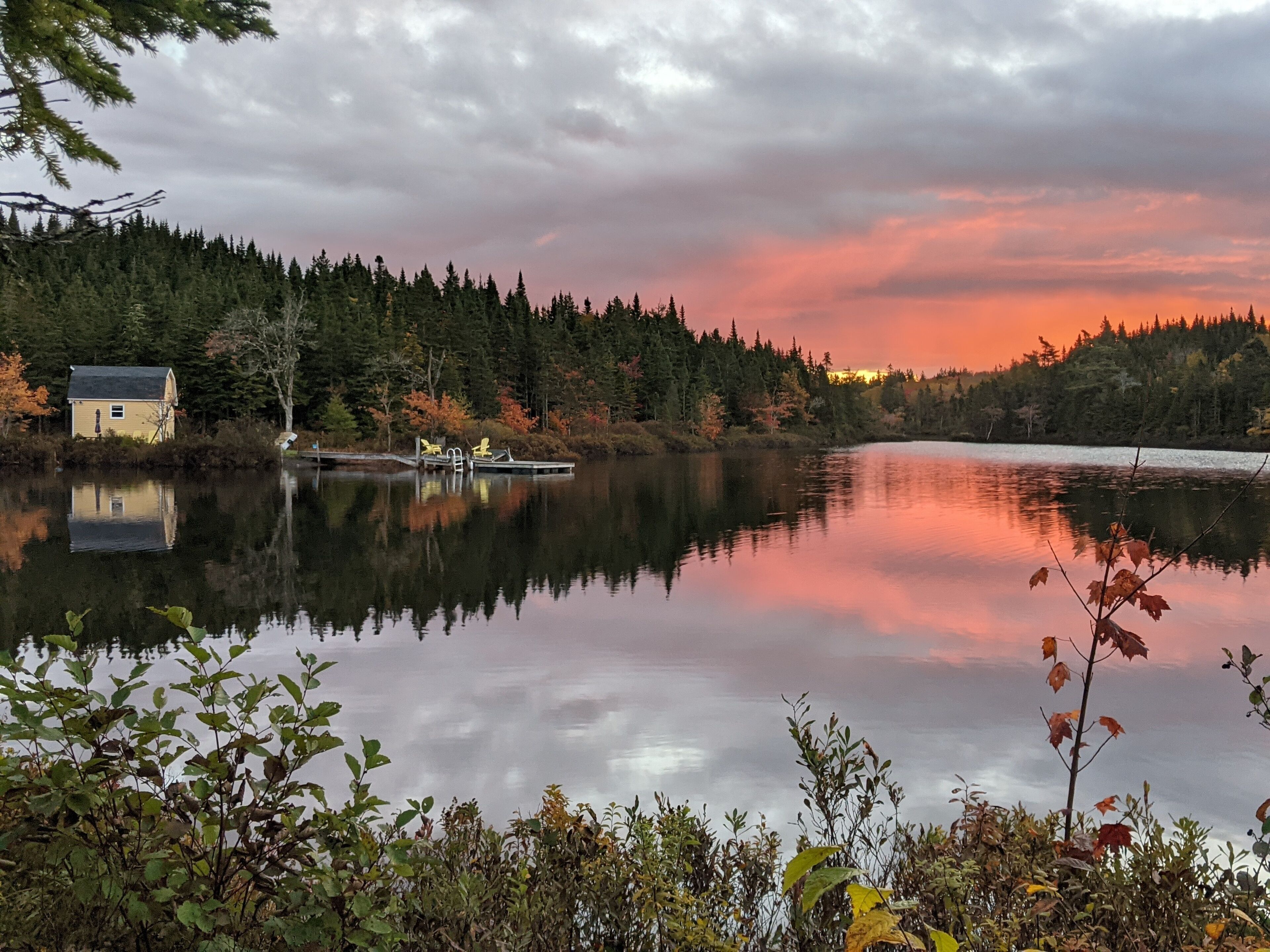 A calm lake with a dock and a house on the left, surrounded by dense forest and a sunset sky.