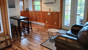 Une pièce avec un plancher en bois, un canapé en cuir, une petite table ronde, un tabouret de bar et un coin cuisine avec un lambris en bois et une fenêtre.