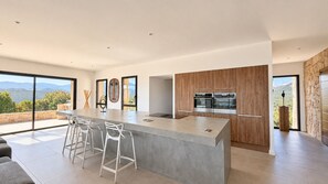A modern kitchen with a large island, bar stools, and a view of the mountains. - Villa Mela di Pinarello (Conca)