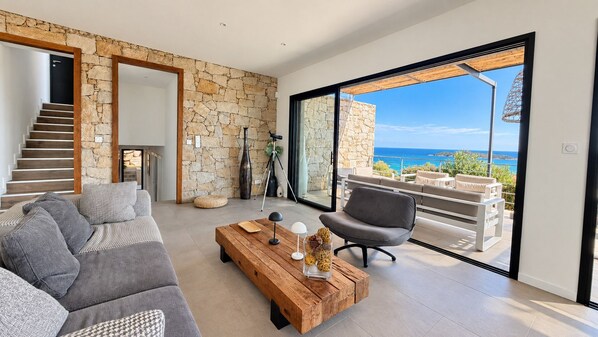 A modern living room with a stone wall, a wooden staircase, a sofa, a coffee table, and a view of the ocean.