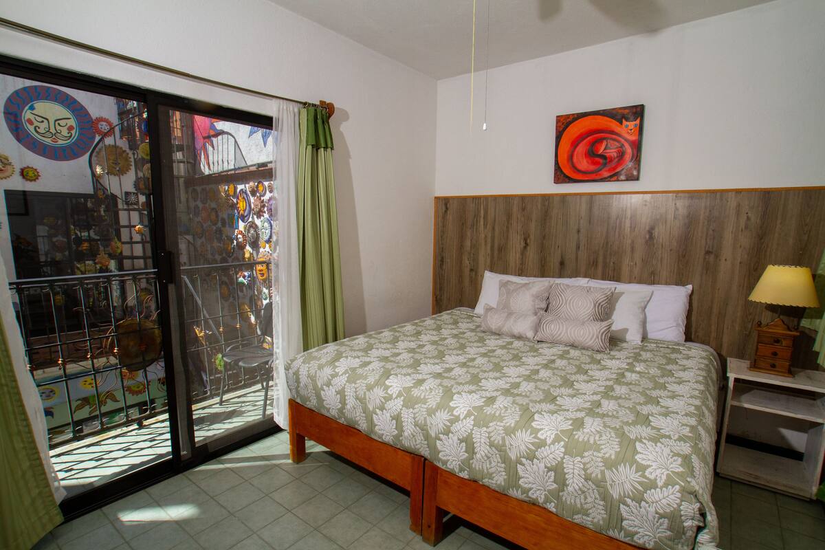 A bedroom with a bed, a wooden headboard, a nightstand, a window with curtains, and a wall with a painting.